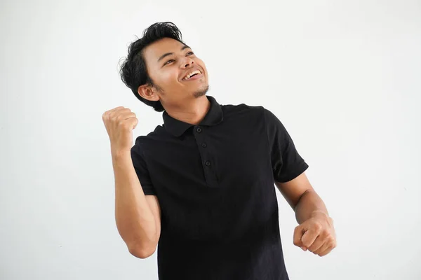 笑顔や幸せな若いアジアの男は ケアフリーで興奮している彼の拳をひねった 白い背景に隔離されたブラックポロTシャツを着用した勝利コンセプト — ストック写真