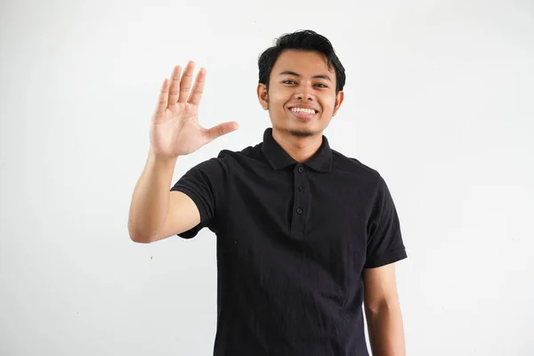 白い背景に隔離された黒いポロTシャツを着ている誰かに挨拶する手を振ると興奮する表情を示す若いアジアの男 — ストック写真