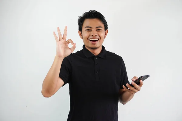 若いアジアの男は自信を持って微笑み 白い背景で隔離された黒いポロTシャツを身に着けている携帯電話を握っている間Okの指の印を与えます — ストック写真
