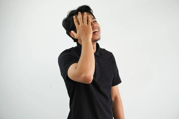 Young asian man posing on a white backdrop having fun covering half of face with palm, wearing black polo t shirt.