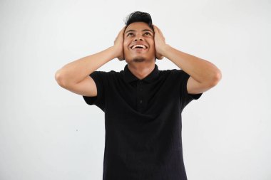 Asyalı mutlu bir genç adam iki elini de kulaklarından tutuyor ve beyaz arka planda siyah polo tişört giyiyor.