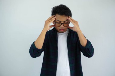 Gözlüklü Asyalı bir genç, beyaz arka planda, başına dokunurken üzgün bir ifade sergiliyor..