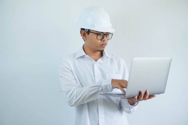 Asyalı ciddi bir mimar, miğfer takıyor ve dizüstü bilgisayarda beyaz arka planda yazıyor.