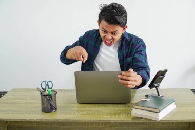 Asyalı kızgın işadamı işyerinde oturmuş bağırıyor ve dizüstü bilgisayarı işaret ediyor.