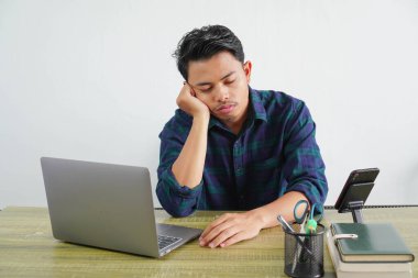 Mavi gömlekli yorgun Asyalı genç adam beyaz arka planda bilgisayar ile ahşap masada çalışıyor. İş hayatının başarısı yaşam tarzı konsepti. ellerin yanaklarında uyu.