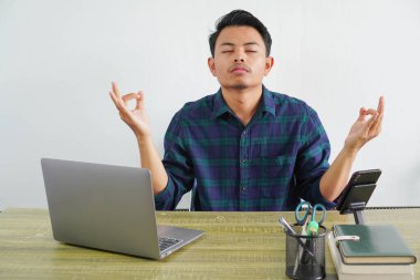 Mavi gömlekli rahat genç Asyalı adam beyaz arka planda bilgisayarlı ahşap masada çalışıyor. İş hayatının başarısı yaşam tarzı konsepti. Yoga jestinde el ele tutuşmak, meditasyon yapmak.