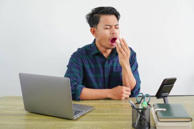 Yorgun Asyalı genç adam ahşap masada bilgisayar başında oturuyor. İş hayatının başarısı yaşam tarzı konsepti. Esniyorlar, ağızlarını eliyle kaplıyorlar.
