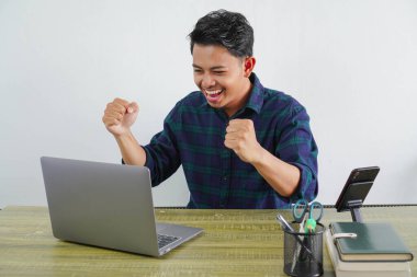 Mutlu Asyalı genç adam ahşap masada bilgisayar başında oturuyor. İş hayatının başarısı yaşam tarzı konsepti. Laptop ekranına bakarken kazanan hareketi yapıyor