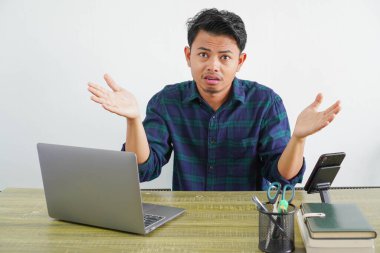 perplexed young asian man sit work at wooden desk with pc laptop. Achievement business career lifestyle concept. spreading hands clipart