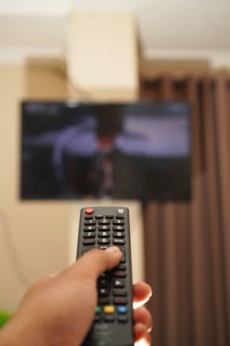 Adamın eli, arka planda televizyon olan uzaktan kumandayı tutuyor. Bakış açısı