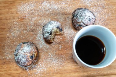 Üç muffin üzerine pudra şekeri serpiştirilmiş, ortadakini ısırılmış, ve ahşap bir masada bir fincan kahve.