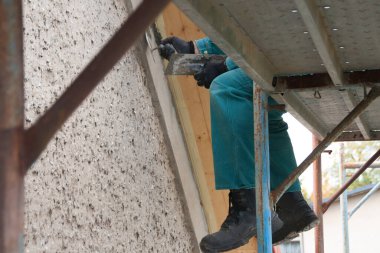 İnşaat işçisinin bacaklarının yakın plan resmi. İskelede oturuyor, güvenli ayakkabılar giyiyor ve çatıdaki bir şeyi mala ve çimentoyla onarıyor.