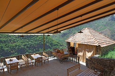 Açık hava restoranında yapılmış ahşap masa ve sandalyeler misafir kabul etmeye hazır, parlak tente çatısı, arka planda orman ve yüksek dağlar, güneşli bir yaz gününde