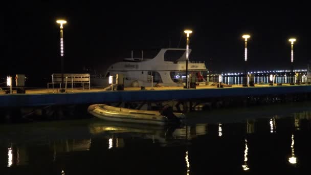 旅游船和充气小艇在夜间停泊在一个灯火通明的码头旁 船尾有沿海灯光 — 图库视频影像