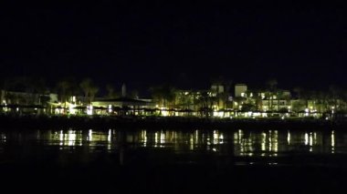 Denizde yansıyan binaları ve palmiyeleri olan gece vakti deniz manzaralı otel, 4k