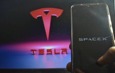 Tesla logosuna karşı SpaceX logosu olan cep telefonunu tutan bir adamın resmi.