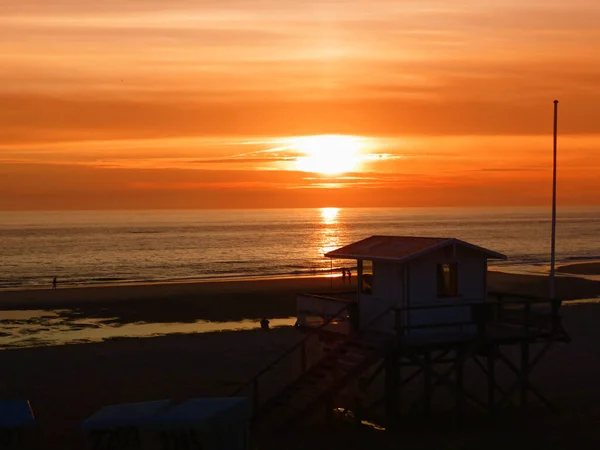 Sylt sahilinde gün batımı, turuncu gökyüzü, ön planda bir sahil evi..