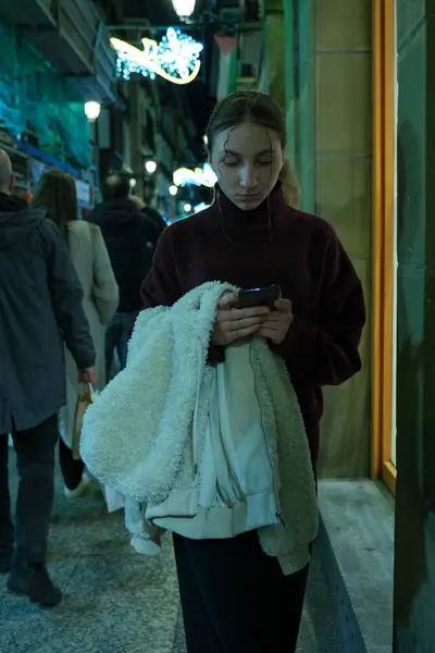 Genç bir kadın yılbaşı pazarında yürürken telefonunu kullanıyor. Akşamları noel pazarında akıllı telefon kullanıyor. Yüksek kalite fotoğraf
