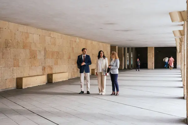 Modern bir binanın koridorunda yürüyen ve konuşan üç iş adamı iş meselelerini tartışıyor.