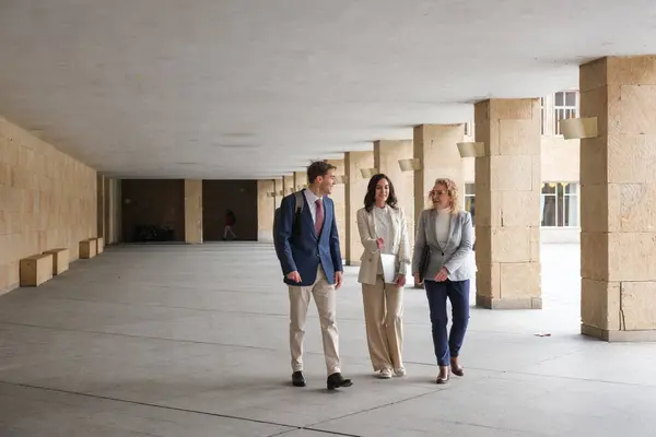 Modern bir binanın koridorunda yürüyen ve konuşan üç iş adamı iş meselelerini tartışıyor.