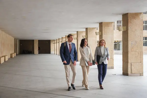 Modern bir binanın koridorunda yürüyen ve konuşan üç iş adamı iş meselelerini tartışıyor.