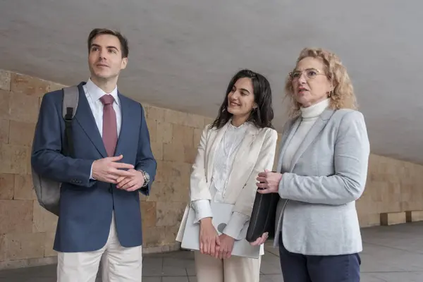 Modern bir binanın koridorunda yürüyen ve konuşan üç iş adamı iş meselelerini tartışıyor.