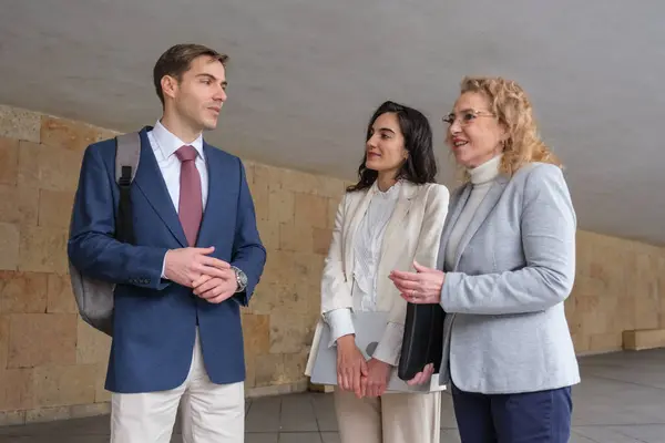 Modern bir binanın koridorunda yürüyen ve konuşan üç iş adamı iş meselelerini tartışıyor.