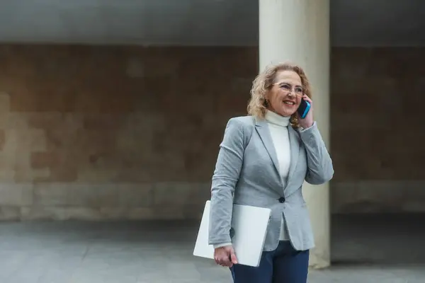 Yetişkin bir kadın, şehir ortamında telefonuyla konuşurken dizüstü bilgisayar tutuyor.