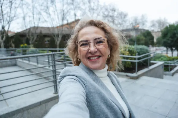 Gülümseyen son sınıf öğrencisi bir kadın, şehir parkında özçekim çekiyor, kendine güven ve neşe gösteriyor.