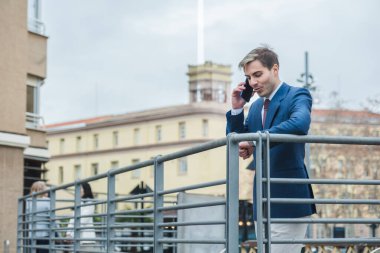 Yetişkin bir iş adamı, şehir ortamında telefonla konuşurken saati kontrol ediyor.