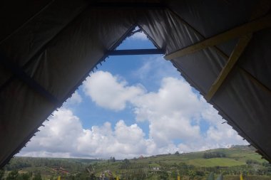 Mavi gökyüzü ve dağlarda beyaz bulutlar olan bir çadır. İçeriden görüntüle.