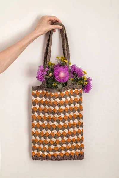 女性の手には紫色の花の入った袋があります 白い背景にイエローホワイトグレーの手作りのチュニジアのかぎ針編みのバッグ 廃棄物ゼロコンセプト — ストック写真