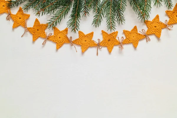 A long yellow crochet garland hanging against a white wall. Christmas handmade star shaped garland and fir tree branches. Christmas decoration. Copy space.