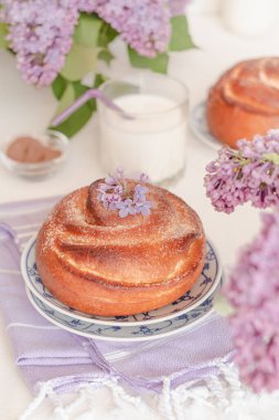 Masada taze pişmiş tarçınlı çörekler ve bir bardak süt ve leylak çiçekleri. Kanelbullar - İsveç tarçını ruloları.