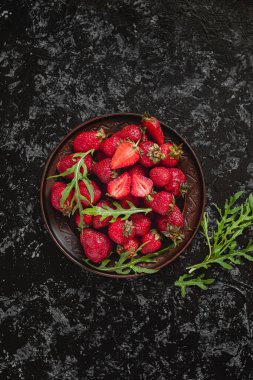 Yuvarlak seramik plaka Wirh taze kırmızı çilek ve koyu arkaplanda roka yaprakları.