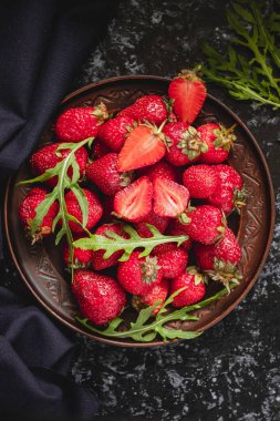 Kahverengi seramik tabakta koyu arkaplanda taze kırmızı çilekler ve roka..