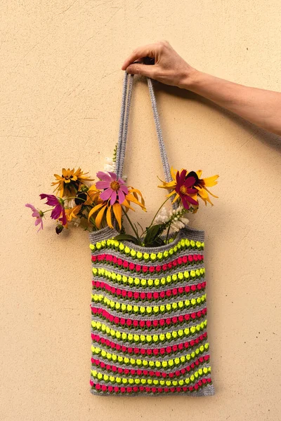 The hand holds a colored knitted bag with summer flowers on a light background. Eko bag crocheted tulip pattern.