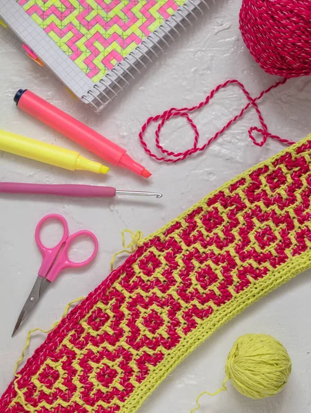 Yellow pink crochet fabric with mosaic pattern. Workplace with crochet fabric, yarn balls, crochet hook, notebook and markers. Handmade concept.