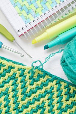 Mozaik desenli sarı yeşil örülmüş doku. Dalgalı örgü dokusu. Tığ işi kumaş, iplik topları, tığ işi kanca, defter ve kalemlerle dolu bir yer..