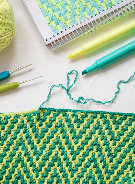 Yellow green crochet fabric with mosaic wave pattern. Workplace with crochet fabric, yarn balls, crochet hook, notebook and markers. Handmade concept.
