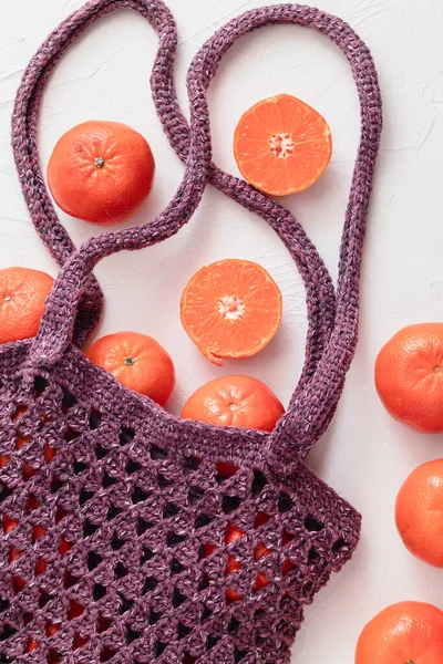 Purple crochet mesh bag with fresh mandarin oranges or tangerines on a white background. Flat lay.
