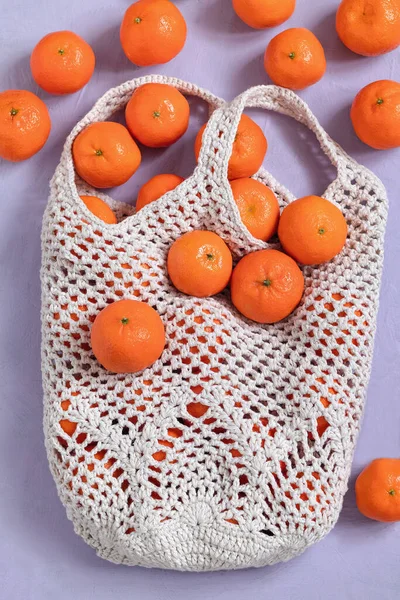 White crochet mesh bag with pineapple pattern. Shopping cotton bag with fresh tangerines on a purple background.