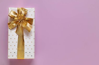 White gift box with golden ribbon and bow on a light purple background. Top view. Copy space.