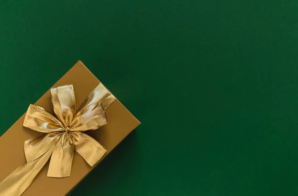 Golden gift box with golden ribbon and bow on a green background. Top view. Copy space.