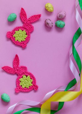 Pink crochet bunnies, colorful eggs and satin ribbon on a pink background. Bright Easter decoration. Top view. Copy space.