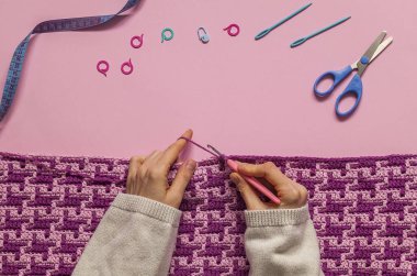 Mor mozaik battaniye ören kadın eller. Pembe arka planda tığ işi kumaş ve tığ işi aletleri. Üst Manzara. Boşluğu kopyala.