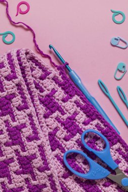 Pembe örgü örme mozaik kumaş, mavi örgü kancası makası. Pembe arka planda iğneler ve işaretler var. Boşluğu kopyala.