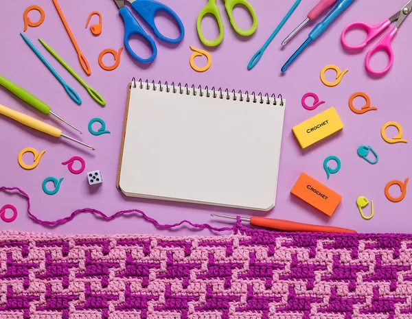 Notepad, crochet fabric and bright colorful crochet tools on a lilac background. Copy space. Top view.