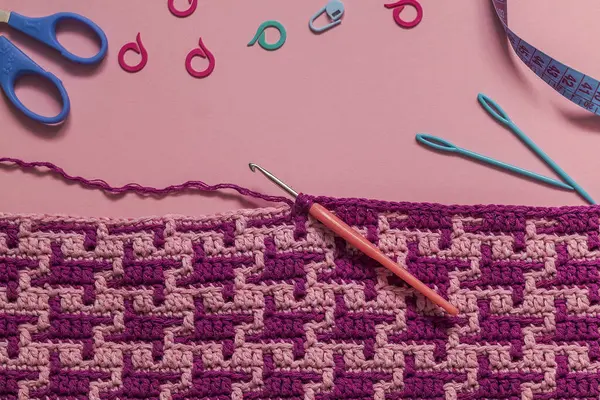 Purple pink crochet fabric with abstract pattern and crochet tools on a pink background. Top view. Copy space.