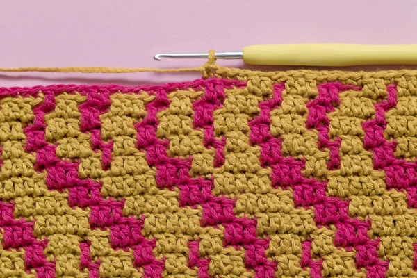Magenta yellow crochet fabric and crochet hook on a pink background. Top view. Copy space.